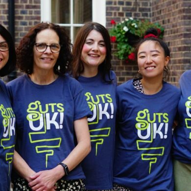 Cherylyn, Melanie, Amy, Leeona and Katie are smiling at the camera wearing Guts UK t-shirts. There is brick wall with white framed windows behind them.