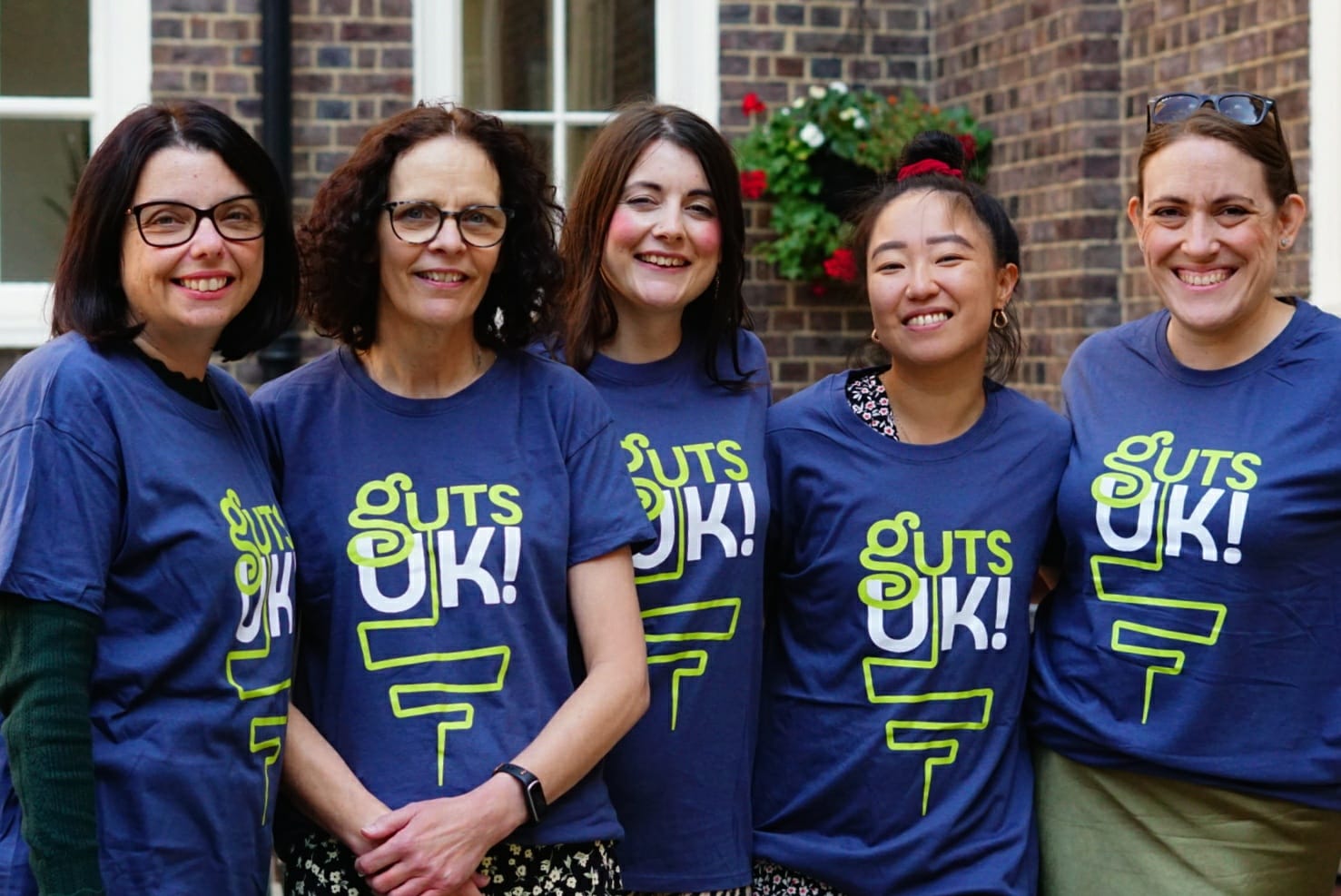 Cherylyn, Melanie, Amy, Leeona and Katie are smiling at the camera wearing Guts UK t-shirts. There is brick wall with white framed windows behind them.