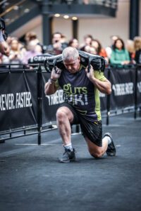 Cornelius has weights on his back and lunges forward. He is wearing a Guts UK t-shirt with black shorts and trainers while completing a race.