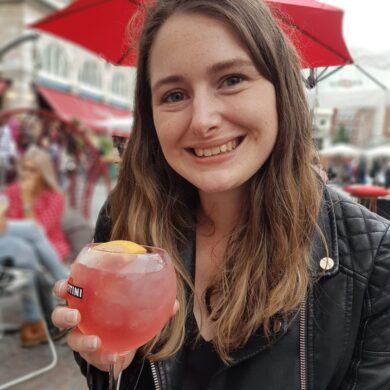 Sian is smiling at the camera in her black jacket. She is sat down at and holding a pink cocktail.