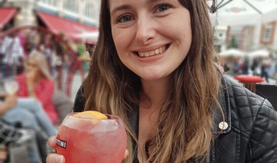 Sian is smiling at the camera in her black jacket. She is sat down at and holding a pink cocktail.