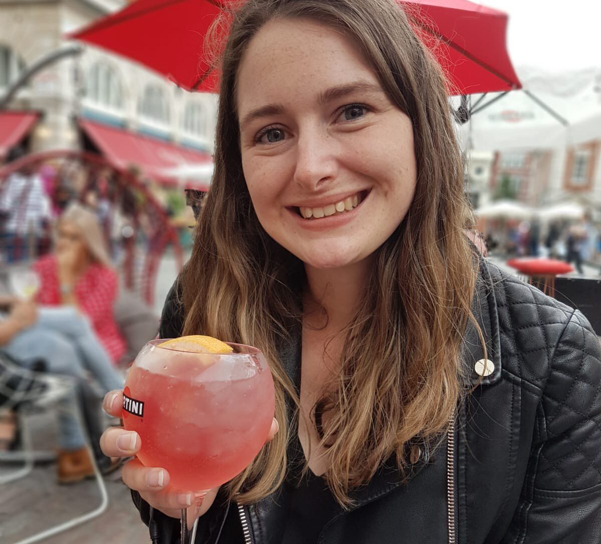 Sian is smiling at the camera in her black jacket. She is sat down at and holding a pink cocktail.
