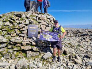 Cornelius is stood up on top of a mountain holding the Guts UK banner. He is wearing the Guts UK t-shirt, a cap and black shorts and smiling at the camera.
