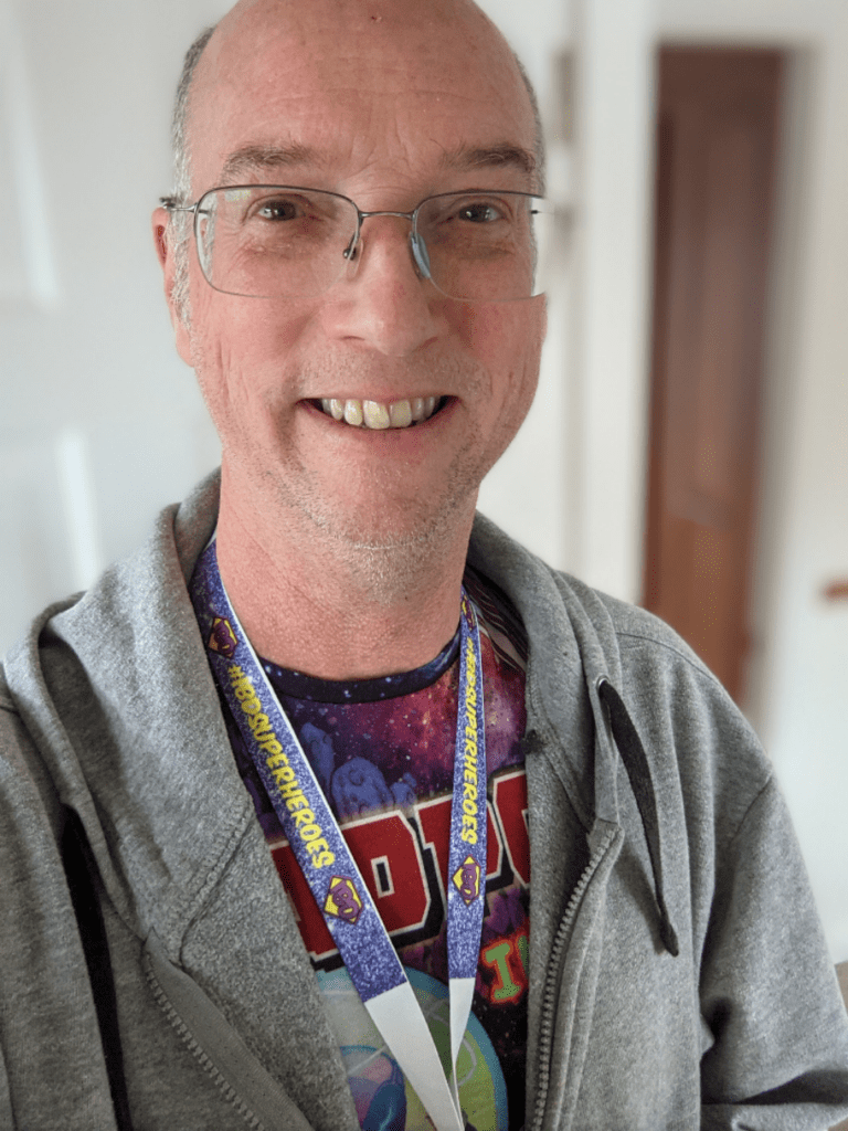Stephen is taking a selfie, smiling at the camera. He wears black framed, rectangular glasses, with curved edges. He has a Deadpool t-shirt on, a grey, zip-up hoodie and an "IBD Superheroes" lanyard around his neck.