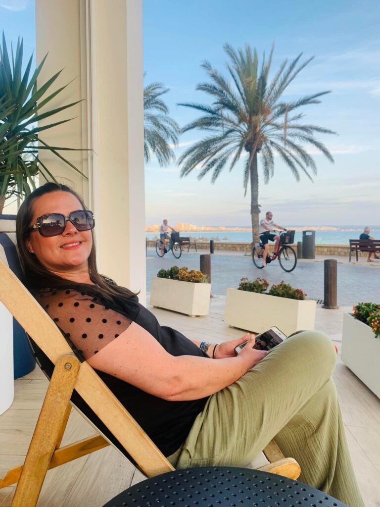 Paula is on holiday in Cyprus, sat in a beach deckchair. She is wearing black, lacey top with polka dot sleeves, khaki linen trousers and black sunglasses. She is smiling at the camera.