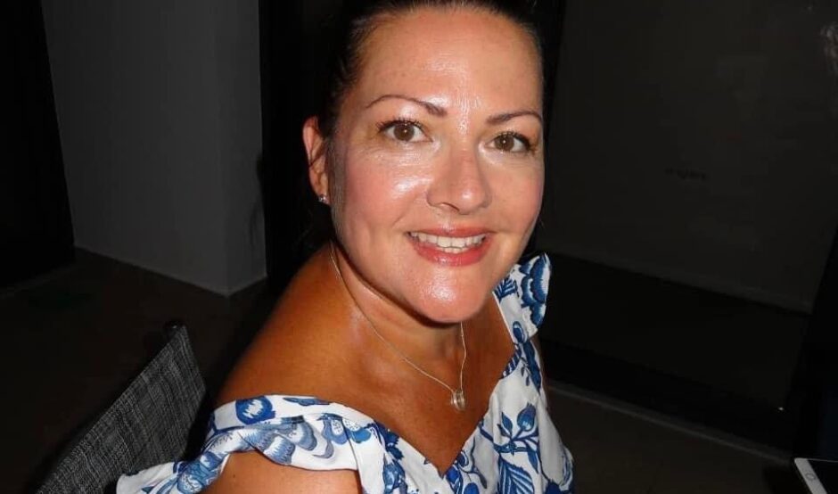 Paula is on holiday in Cyprus, ready for a night out. The photo is taken from slightly above her, and she smiles at the camera. She is wearing a white and blue floral dress, with frilly straps over her shoulders.