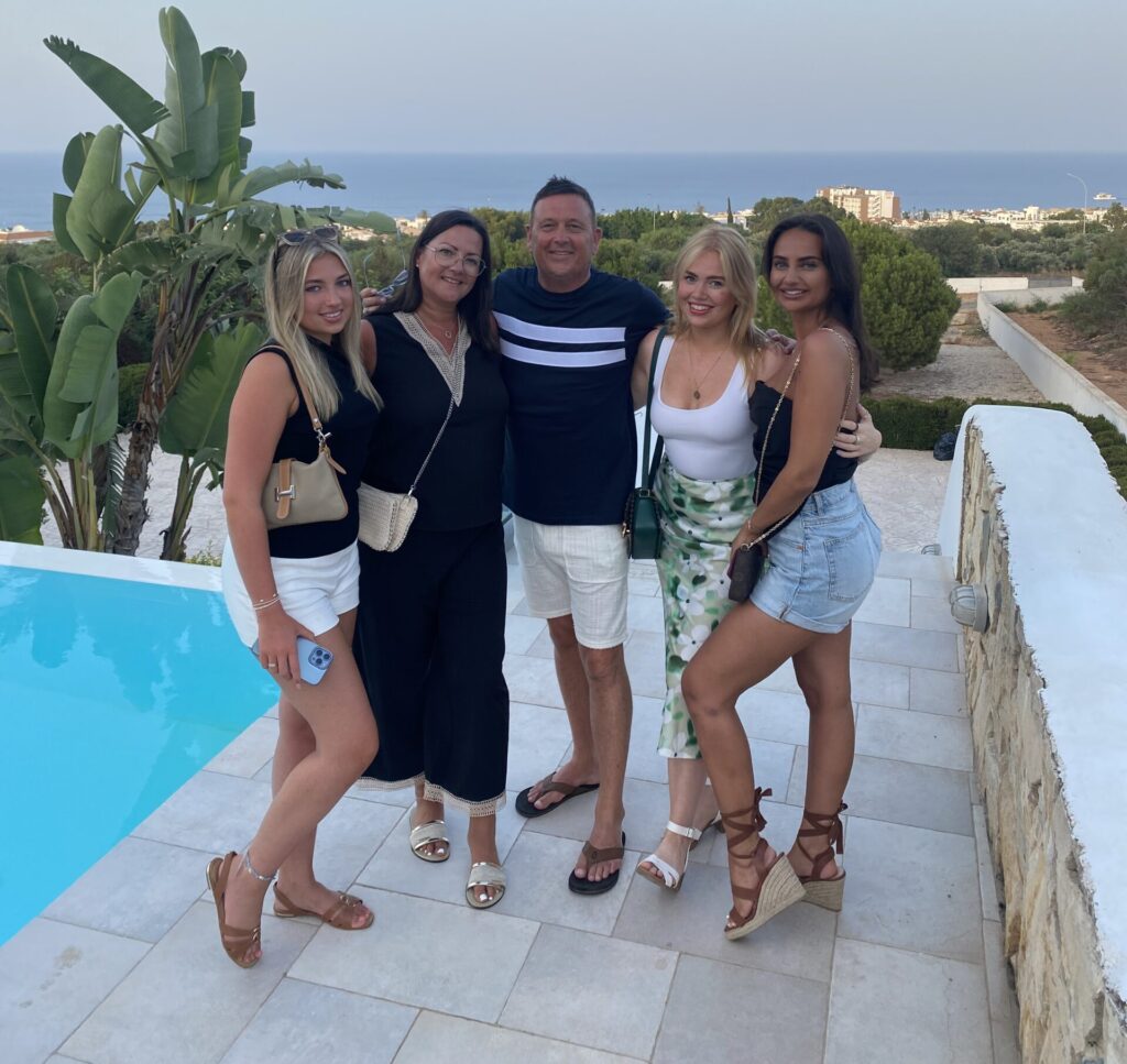 Paula is on holiday in Cyprus with her family. She is smiling at the camera and wears a ankle-length black dress, with pale sandals and a pale cross-body bag. There is a swimming pool to her left, filled with bright blue water, and you can see the scenery of Cyprus and the sea in the background.