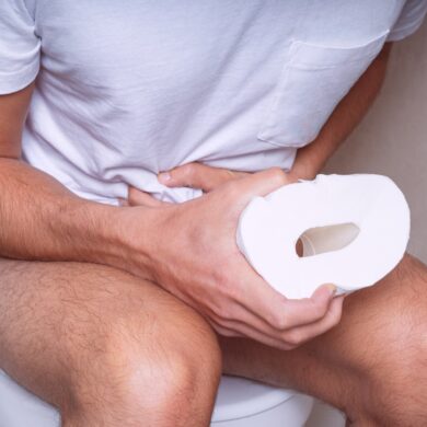 A person is sat on the toilet. They are wearing a white t-shirt. They are holding a roll of toilet paper in one hand and clutching their stomach in the other.