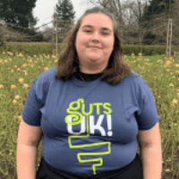 Frankie has short, shoulder length brown hair. She is wearing a navy blue t-shirt that says 'Guts UK' and shows an illustration of the outline of a digestive system. She is standing outside and is smiling at the camera. Behind her, there is a grass area with yellow flowers.
