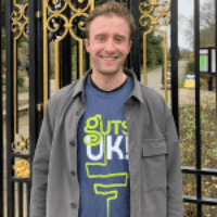 Freddy has short, light hair. He is stood outside, smiling at the camera. Behind him are black and gold railings and grass. He is wearing a blue Guts UK t-shirt with a grey jacket with buttons over the top.