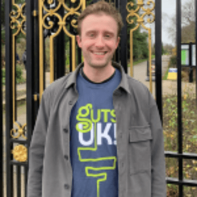 Freddy has short, light hair. He is stood outside, smiling at the camera. Behind him are black and gold railings and grass. He is wearing a blue Guts UK t-shirt with a grey jacket with buttons over the top.