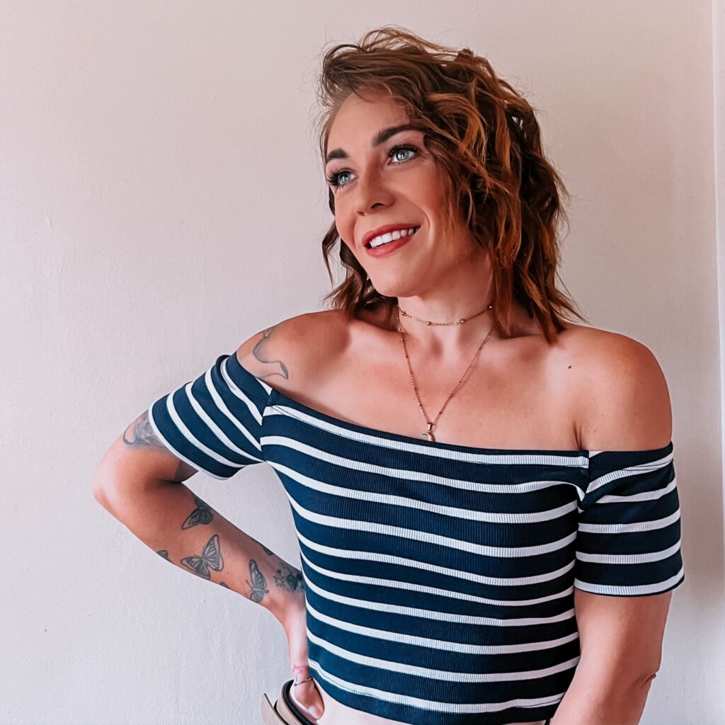 Rachel wears a navy and white horizontally striped, short-sleeved, off the shoulder top, and smiles as she looks left. She has short, brown, wavy hair, blue eyes, and also wears a gold necklace. She has butterfly tattoos on her left arm and places her left hand on her hip. Her right arm rests straight down the side of her body.