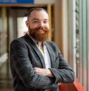 A portrait photograph of Dr Benjamin Sharpe. He is standing up with his arms folded in front of him and is smiling at the camera.