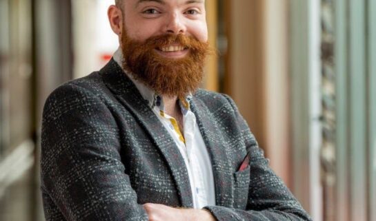 A portrait photograph of Dr Benjamin Sharpe. He is standing up with his arms folded in front of him and is smiling at the camera.