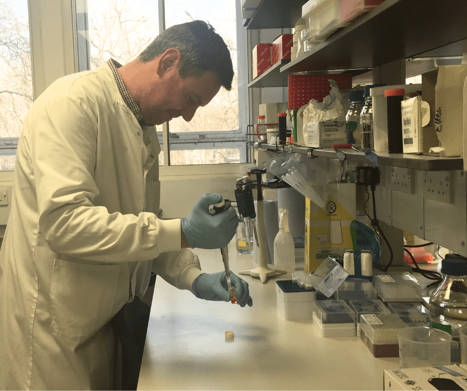 Dr McCann is in a research laboratory. He has a pipette in his hand and is putting a sample into a container. He wears a white laboratory coat and blue medical gloves.