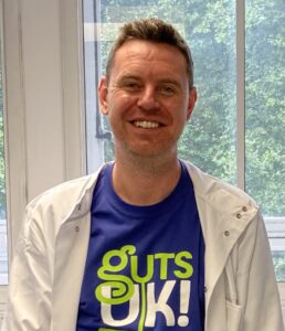 Dr Conor McCann wears a blue Guts UK t-shirt, underneath a white lab coat and smiles at the camera.