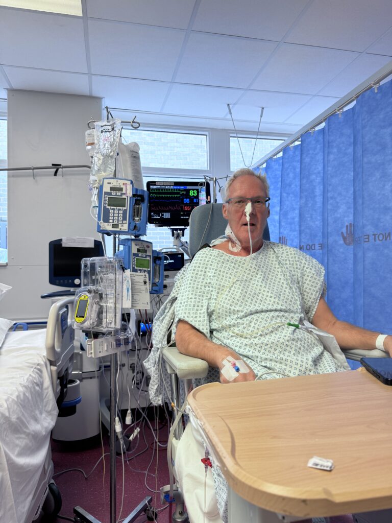 Graham is sat in a hospital chair on a ward. He is wearing a hospital gown and has a tube into his nose and various machines around him.
