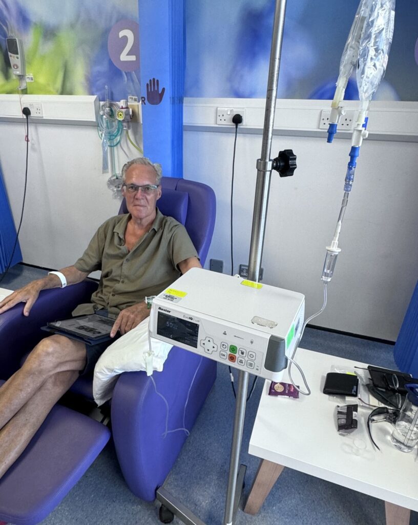 Graham is sat in a blue hospital chair, receiving chemotherapy treatment via a line into his arm.