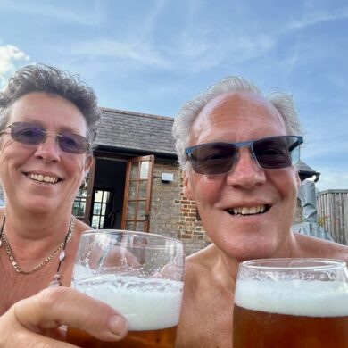 Sarah and Graham are sat enjoying a pint of beer each outside. They are both wearing sunglasses and the sky is blue.