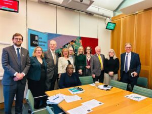 Members of the All-Party Parliamentary Group (APPG) on Less Survivable Cancers meet. They are stood and sat around a boardroom table. Papers can be seen on the table.