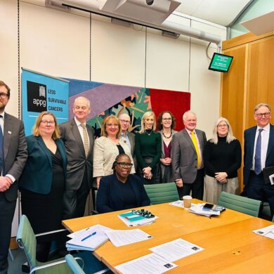 Members of the All-Party Parliamentary Group (APPG) on Less Survivable Cancers meet. They are stood and sat around a boardroom table. Papers can be seen on the table.