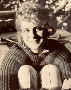 A black and white image of Claire sat down hugging her legs. She is smiling at the camera and wearing a jumper with jeans.