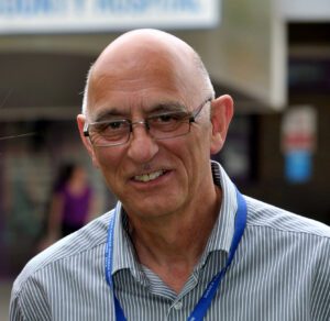 Dave is wearing glasses, a striped blue shirt and a blue lanyard. He is smiling at the camera. The background is blurred out. Only his head and shoulders are visible in the image.