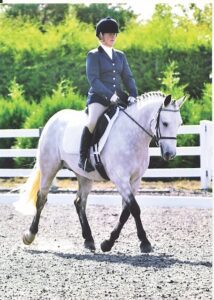 Claire is riding on a white horse in her equestrian clothing. Green bushes can be seen behind them.