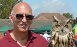 Dave is smiling at the camera. He is wearing shades and a red shirt. There is a great horned owl next to him. The sky is blue with clouds. Some brick buildings and tents re seen behind them.