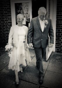 A black and white photo of Claire and her husband walking towards the camera smiling. Claire is wearing a white knee length dress and her husband is wearing a suit. 