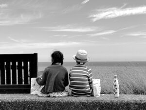 A black and white image of two children sat next to a bench and looking onwards to the sea. 