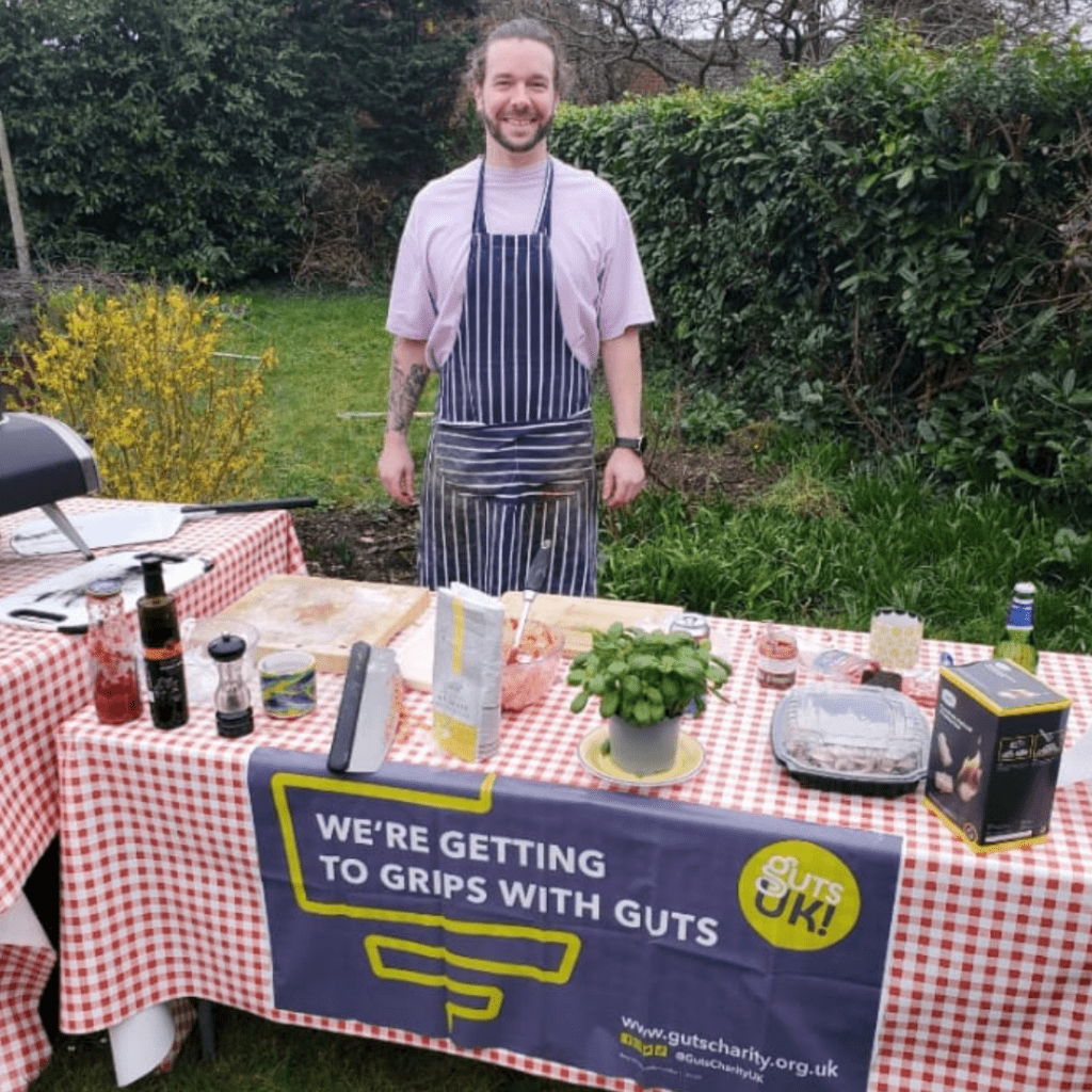 Damien is smiling at the camera and wears a blue and white striped apron. He has a table in front of him, outdoors, next to a pizza oven with various condiments and cutlery for making pizzas.