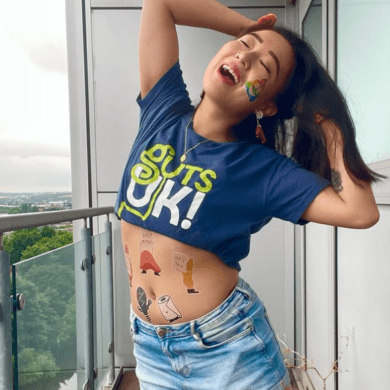 Leeona is tilting her head back, smiling, with her hands in her hair. She wears a Guts UK t-shirt and temporary #GUTSelfie tattoos are on her stomach.