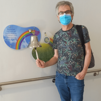 Steve stands in a hospital corridor, ringing a bell. There is a sign behind it with a rainbow. He is wearing jeans and a shirt. He has a mask covering his face.