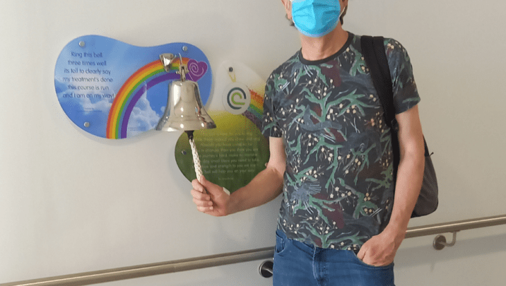 Steve stands in a hospital corridor, ringing a bell. There is a sign behind it with a rainbow. He is wearing jeans and a shirt. He has a mask covering his face.
