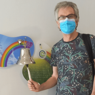 Steve stands in a hospital corridor, ringing a bell. There is a sign behind it with a rainbow. He is wearing jeans and a shirt. He has a mask covering his face.