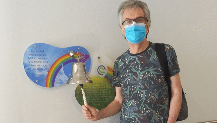 Steve stands in a hospital corridor, ringing a bell. There is a sign behind it with a rainbow. He is wearing jeans and a shirt. He has a mask covering his face.
