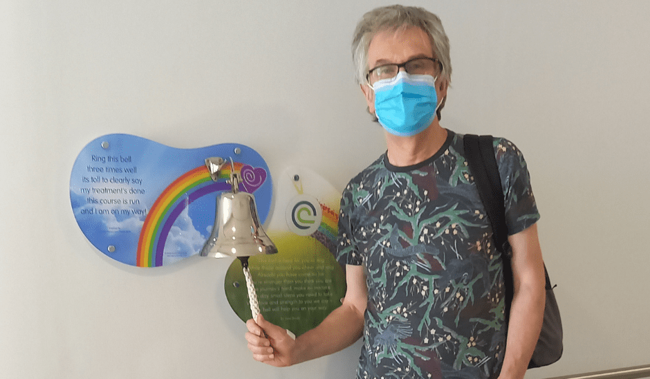 Steve stands in a hospital corridor, ringing a bell. There is a sign behind it with a rainbow. He is wearing jeans and a shirt. He has a mask covering his face.
