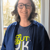 Lynn is wearing a navy Guts UK t-shirt and a denim shirt, opened. She is smiling at the camera and wears round glasses.