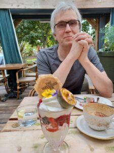 Steve is wearing glasses and a blue tshirt. He is sat at a table, his hands clasped together in front of him, with elbows resting on the table. There is a coffee and an ice cream on the table in front of him.