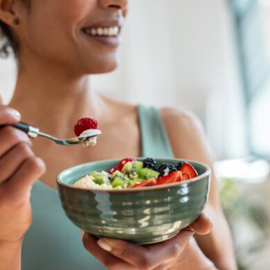 A woman is holding a green bowl and a spoon. She is eating fruit and yogurt out of a bowl. She is wearing a blue vest top and is smiling.