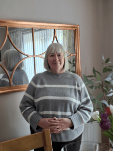Diane is stood up, smiling at the camera. She has short blonde hair and is wearing a grey jumper with white stripes and black trousers.
