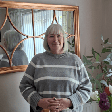 Diane is stood up, smiling at the camera. She has short blonde hair and is wearing a grey jumper with white stripes and black trousers.