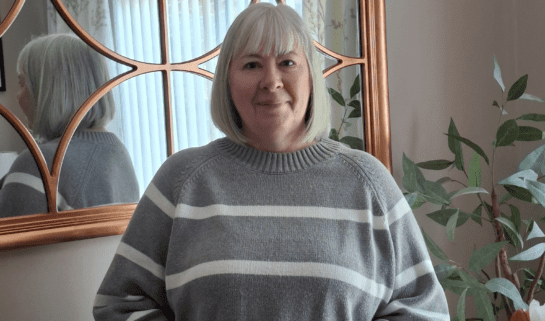 Diane is stood up, smiling at the camera. She has short blonde hair and is wearing a grey jumper with white stripes and black trousers.