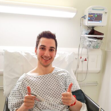 Ziyad is sitting up in a hospital bed, wearing a hospital gown. A medical monitor is beside him. He’s smiling at the camera with both thumbs up.