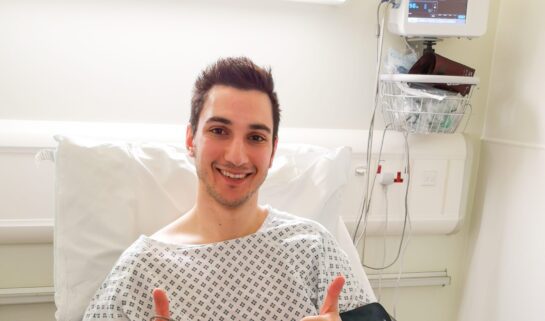Ziyad is sitting up in a hospital bed, wearing a hospital gown. A medical monitor is beside him. He’s smiling at the camera with both thumbs up.