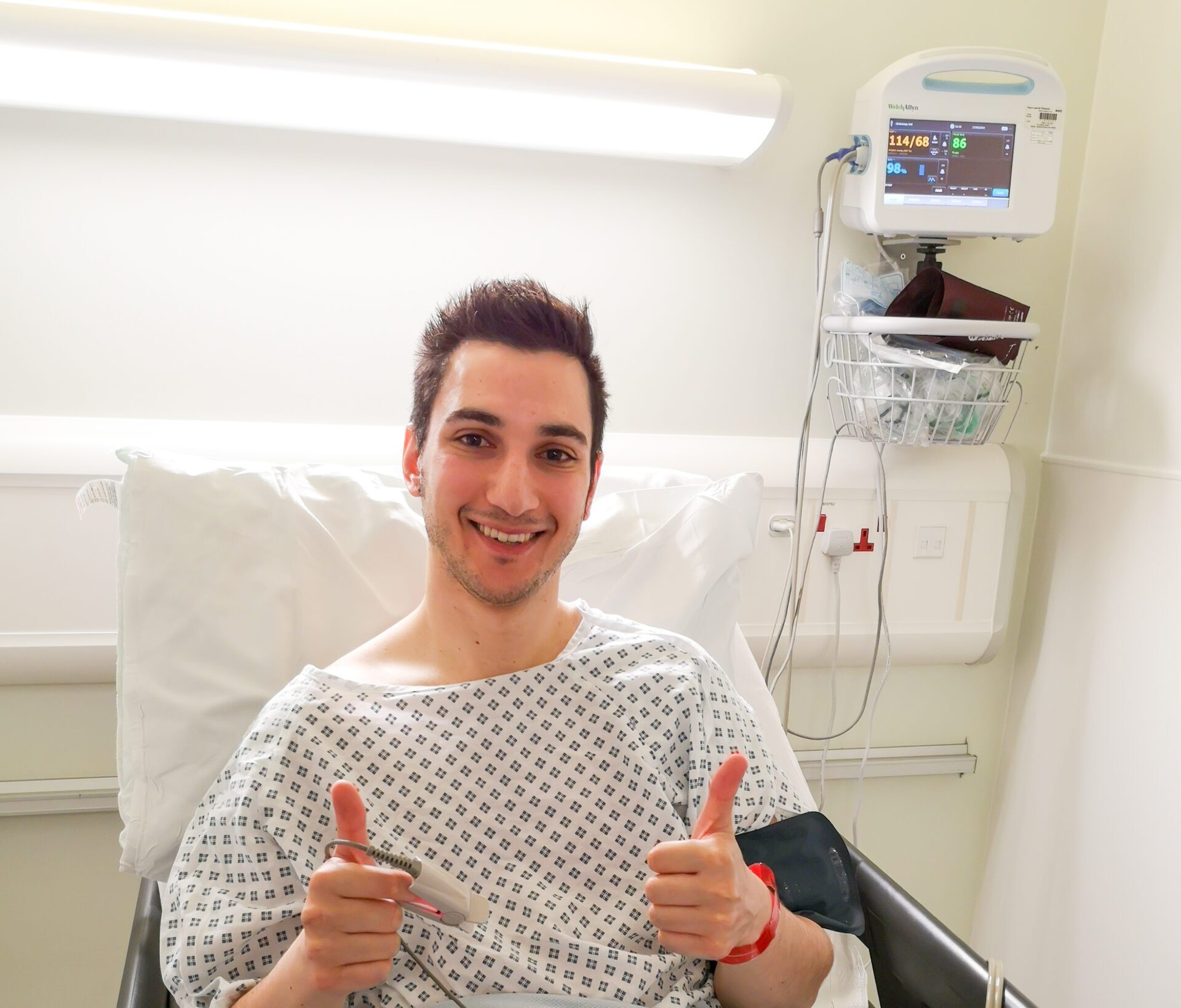 Ziyad is sitting up in a hospital bed, wearing a hospital gown. A medical monitor is beside him. He’s smiling at the camera with both thumbs up.