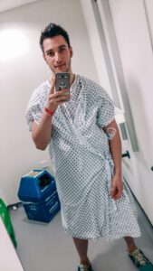 Ziyad stands in a hospital room, wearing a hospital gown and smiling as he takes a mirror selfie. 