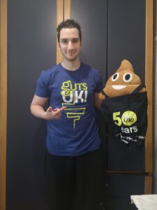 Ziyad is standing in front of a black wall, wearing a Guts UK t-shirt and black trousers. He’s holding a Guts UK 50 years tote bag with a poo emoji cushion resting on top. He’s smiling at the camera and pointing at the poo emoji with his other hand. 