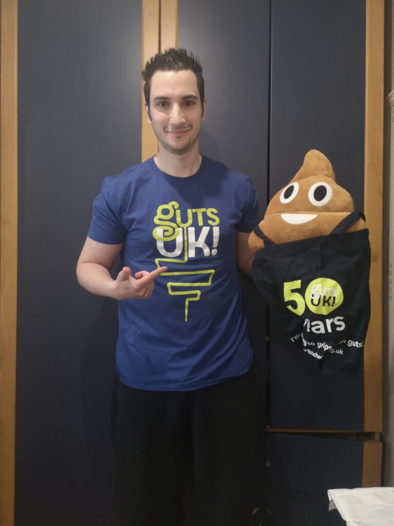Ziyad stands in front of a dark blue wardrobe, smiling and pointing toward the logo on his t-shirt. He is wearing a blue t-shirt that says "GUTS UK!" in lime green and white letters, with a stylized line drawing of a digestive tract underneath. Under his other arm, he holds a large plush toy shaped like the smiling poop emoji, which is tucked into a black tote bag also featuring the "GUTS UK!" logo and the number 50.